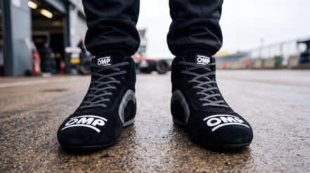Close up at ground level of a racing driver stood in a pit lane wearing black OMP suede race boots, the OMP wordmark across both toe boxes