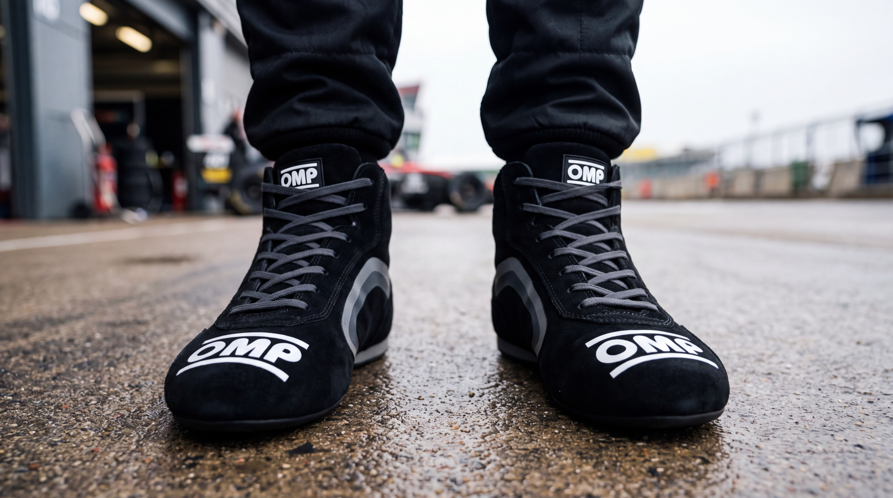 Close up at ground level of a racing driver stood in a pit lane wearing black OMP suede race boots, the OMP wordmark across both toe boxes