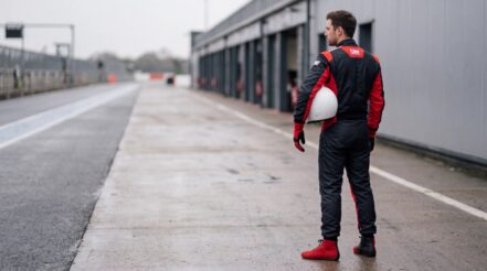 Racing driver in the OMP Tecnica Evo race suit in Black & Red looking down a circuit pit lane, plain white helmet under one arm