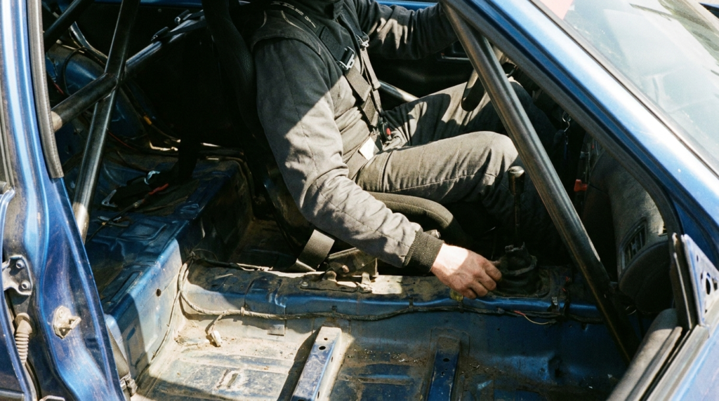Driver harnessed into a bucket seat in a stripped competition car reaching down beside the seat