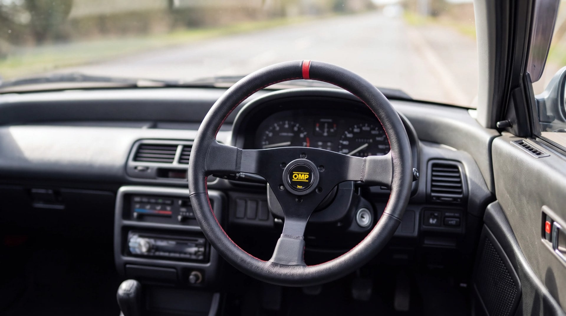 Interior view of a right-hand-drive Honda Civic from the late 1980s fitted with an OMP steering wheel and boss kit, viewed from the driver's seat