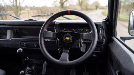 Land Rover Defender 90 interior with an OMP steering wheel boss kit fitted, RHD layout, original Defender dashboard, rural English countryside through the windscreen