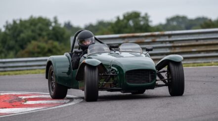 Open cockpit two seater club racing car mid corner on a British circuit, the driver wearing a plain full face crash helmet with the visor down