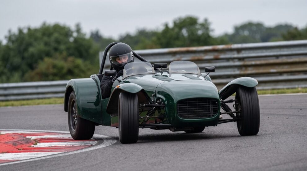 Open cockpit two seater club racing car mid corner on a British circuit, the driver wearing a plain full face crash helmet with the visor down