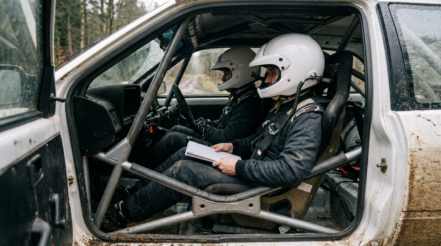 Driver and co-driver strapped into a rally car wearing plain white open face helmets with intercom leads and pace notes