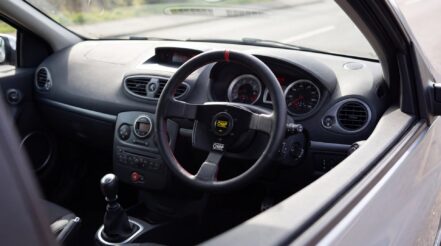 OMP three-spoke steering wheel on a right-hand-drive Renault Clio III RS, driver's door visible on the right, gear lever to the left, dashboard beyond