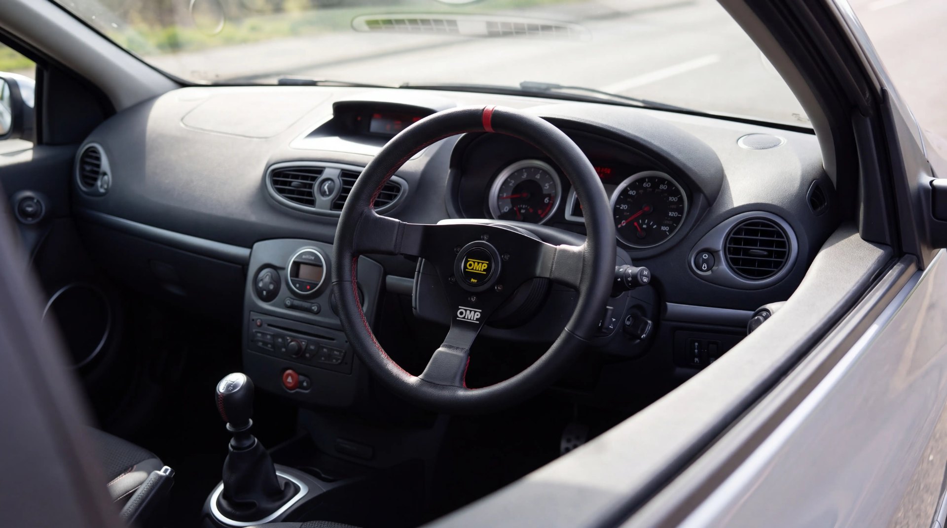 OMP three-spoke steering wheel on a right-hand-drive Renault Clio III RS, driver's door visible on the right, gear lever to the left, dashboard beyond