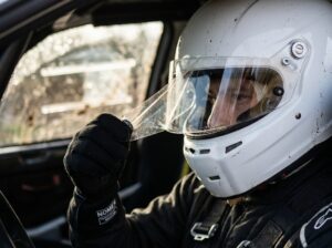 Gloved hand peeling a dirty transparent tear-off strip from the clear visor of a white rally helmet