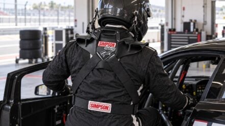 Helmeted racing driver wearing a Simpson Hybrid Sport head and neck restraint beside a race car