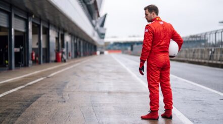 Racing driver in the OMP Sport race suit in Red looking down a circuit pit lane, plain white helmet under one arm