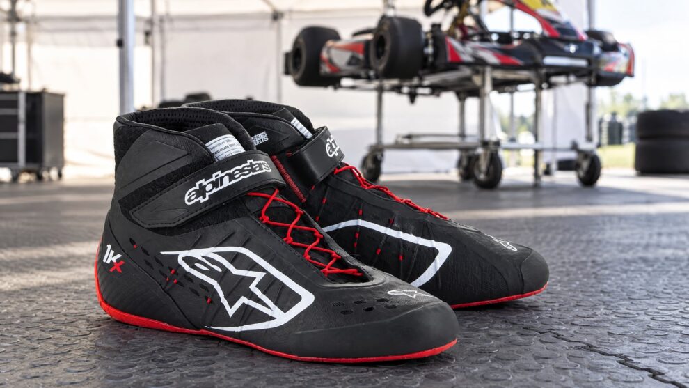A pair of black and red Alpinestars Tech 1-KX kart boots on a paddock floor, with a kart on a stand behind