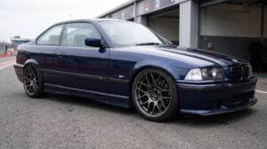A BMW E36 coupe slammed on coilovers in the pit lane, dark aftermarket wheels with open hubs