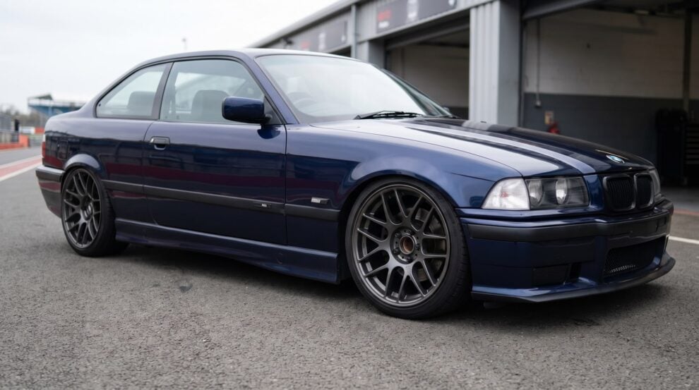 A BMW E36 coupe slammed on coilovers in the pit lane, dark aftermarket wheels with open hubs