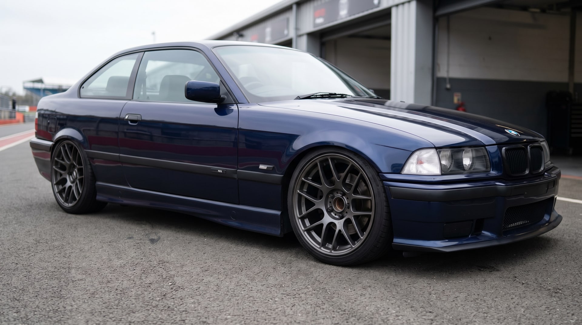 A BMW E36 coupe slammed on coilovers in the pit lane, dark aftermarket wheels with open hubs