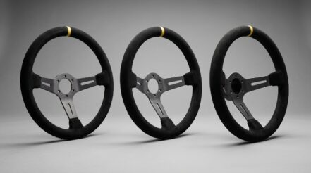 Three deep dish racing steering wheels side by side, each with its suede rim standing well forward of the mounting hub and the spokes swept back into the dish