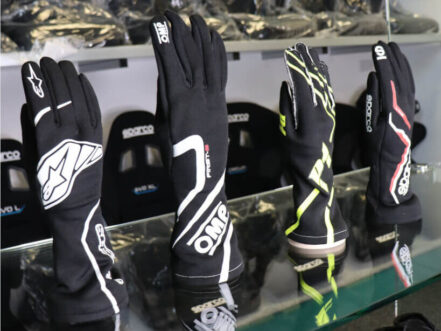 Four FIA race gloves from Alpinestars, OMP, P1 and Sparco standing on a glass shelf in the GSM Performance showroom