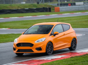 Orange Ford Fiesta ST at a British race circuit apex with helmeted driver, red and white kerbing visible, overcast trackday light