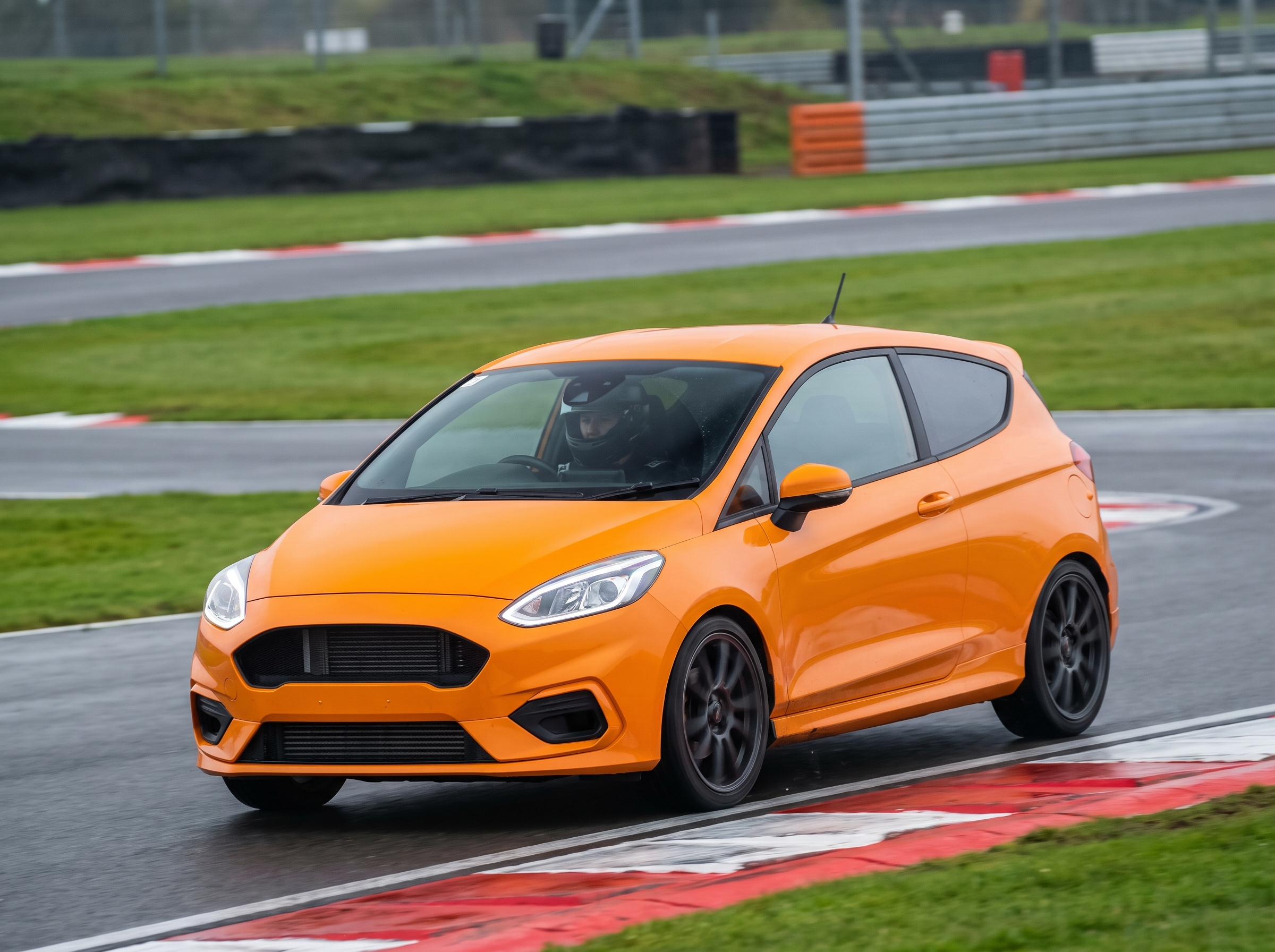Orange Ford Fiesta ST at a British race circuit apex with helmeted driver, red and white kerbing visible, overcast trackday light