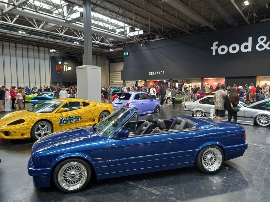 Dark blue BMW E30 convertible on BBS RS wheels in front of a yellow wrapped Ferrari 360 and a purple Abarth 500 in the NEC hall