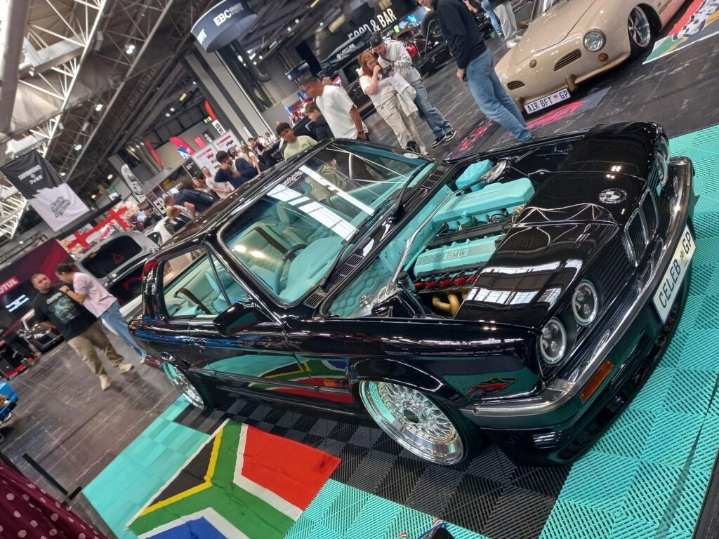 Black BMW E30 with a South African flag livery, teal painted engine and BBS wheels on a teal tiled stand