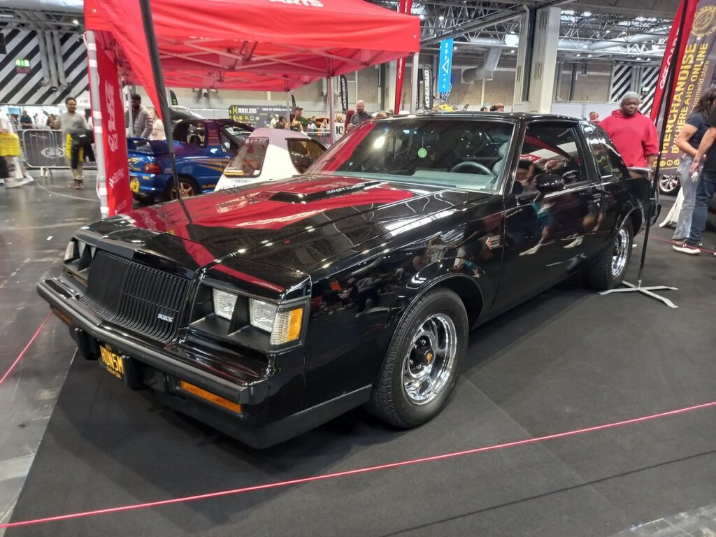 Black 1987 Buick Grand National turbo coupe on chrome wheels under a red gazebo at Gravity Show 2026