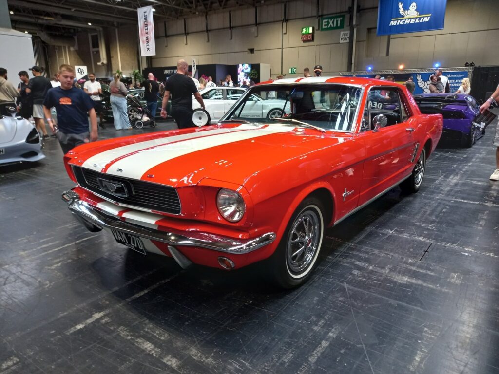 Red 1966 Ford Mustang coupe with white racing stripes and whitewall tyres, a white McLaren and a purple Lamborghini behind