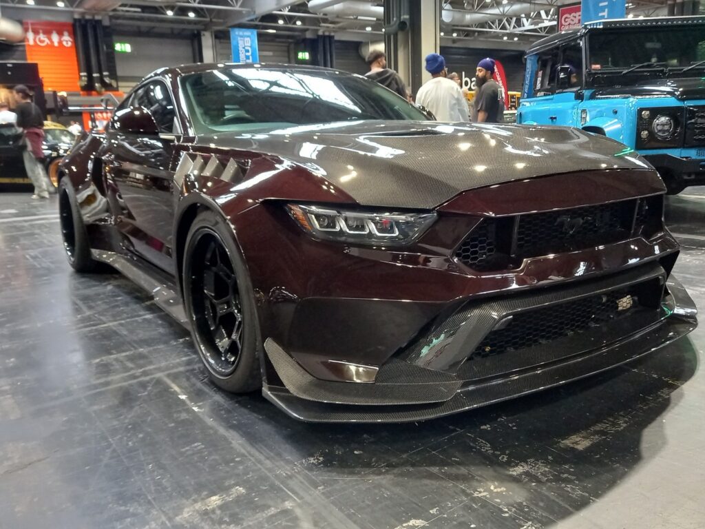 Black cherry Ford Mustang S650 wide-body with a carbon bonnet and splitter, from the front, a blue Land Rover Defender behind
