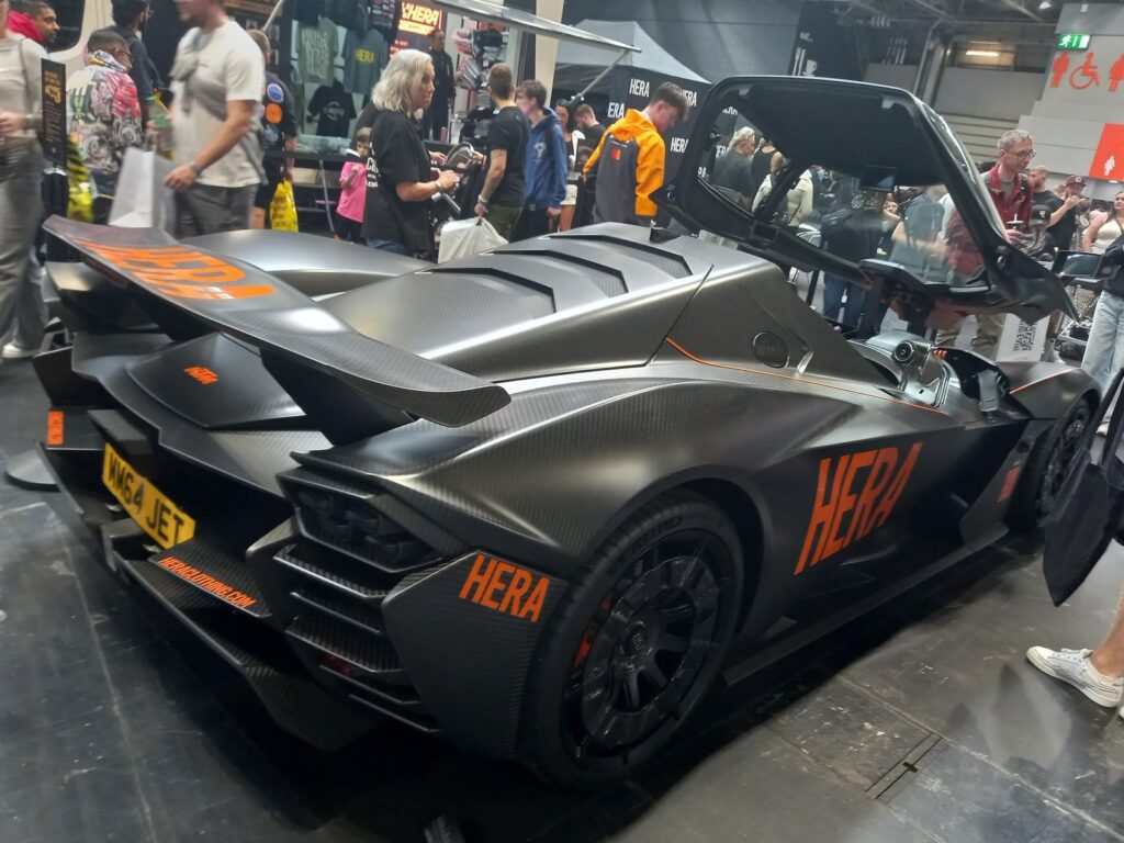 Matte carbon KTM X-Bow GT-XR with its canopy open and orange HERA branding, rear three quarter view