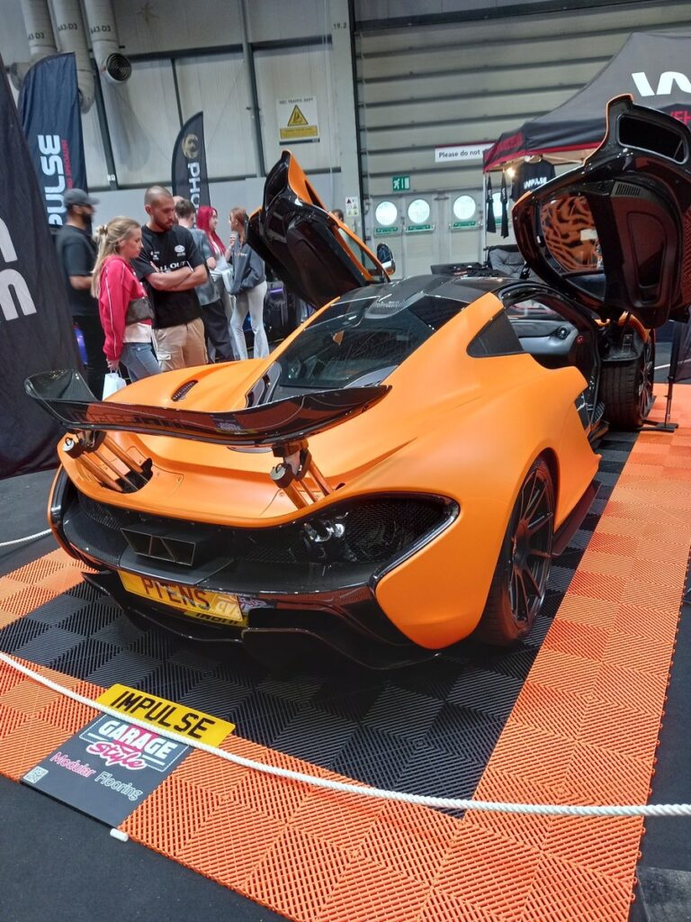 Matte orange McLaren P1 with both doors raised and the rear wing extended, on orange modular flooring