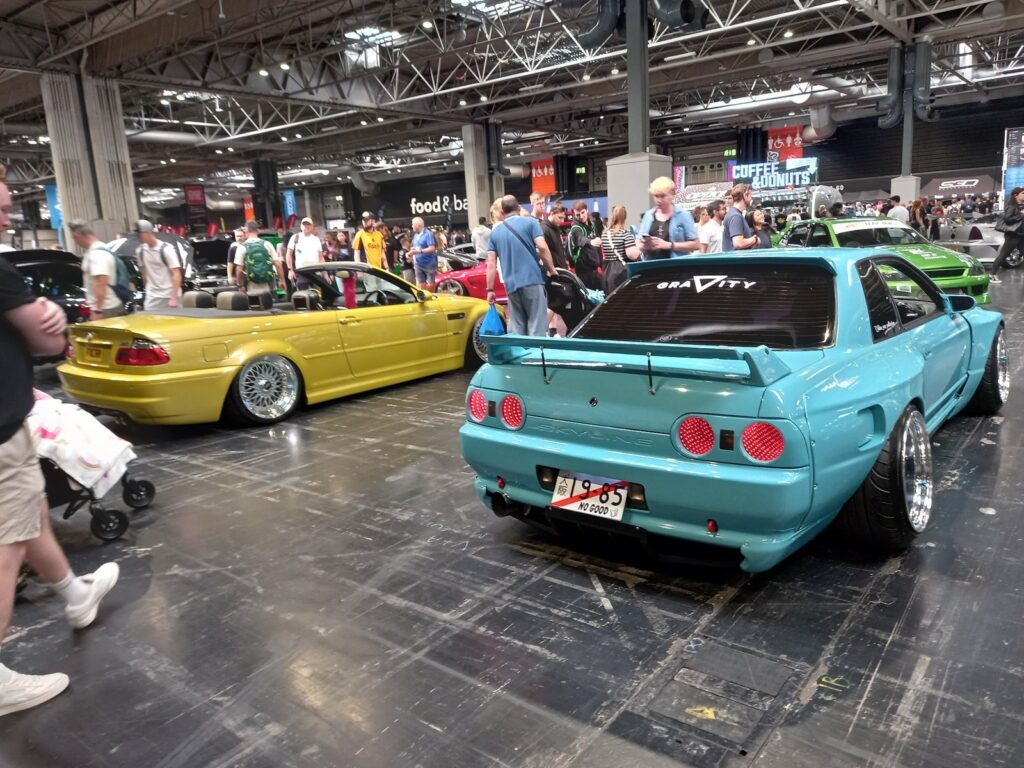 Teal Nissan Skyline R32 next to a yellow BMW E46 M3 convertible on BBS wheels in the NEC hall