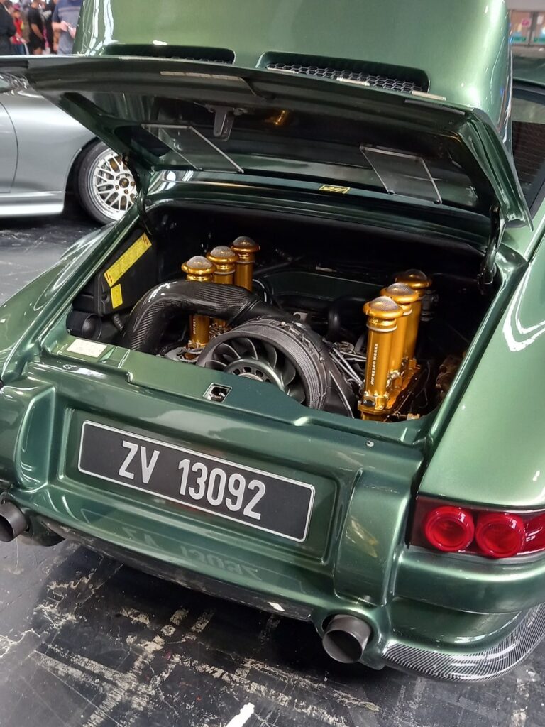Engine bay of a dark green Porsche 911 restomod with gold individual throttle body trumpets and a carbon airbox