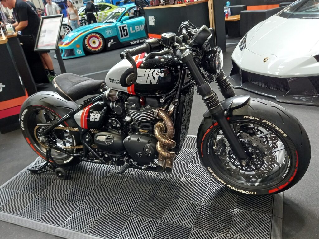 Custom Triumph Bobber by Thornton Hundred on the K and N stand, with a Le Mans style Porsche and a white Lamborghini behind