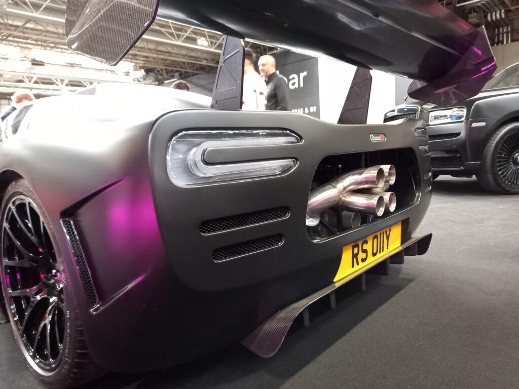 Rear of an Ultima RS in colour-shift purple with its quad exhaust, carbon diffuser and towering rear wing, a Rolls-Royce Cullinan behind