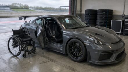 A grey GT4 race car with the driver door open in a pit garage, a wheelchair parked beside it and tyres stacked behind