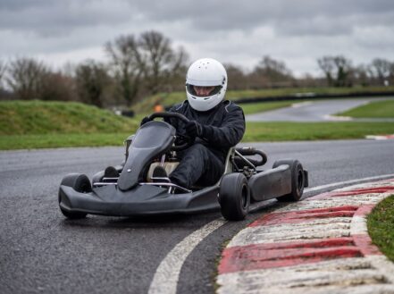 Racing kart at a British outdoor karting circuit, driver in plain helmet with gloved hands on the steering wheel at an apex with red and white kerbing
