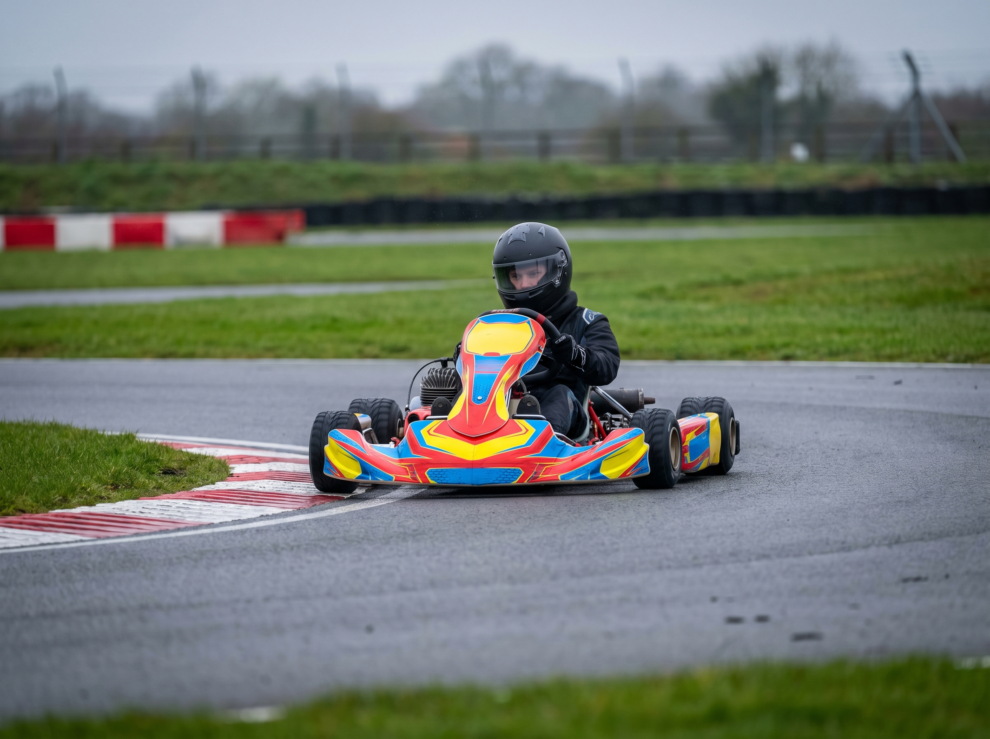 Racing kart at a British outdoor karting circuit, driver in full face helmet at an apex on smooth tarmac, overcast light, kart and driver fully in frame