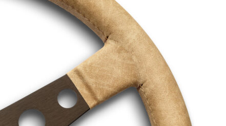 Close up of the MOMO Prototipo Desert steering wheel where the dark brown anodised spoke meets the Sahara vintage leather rim, with the parallel sand stitching visible along the rim
