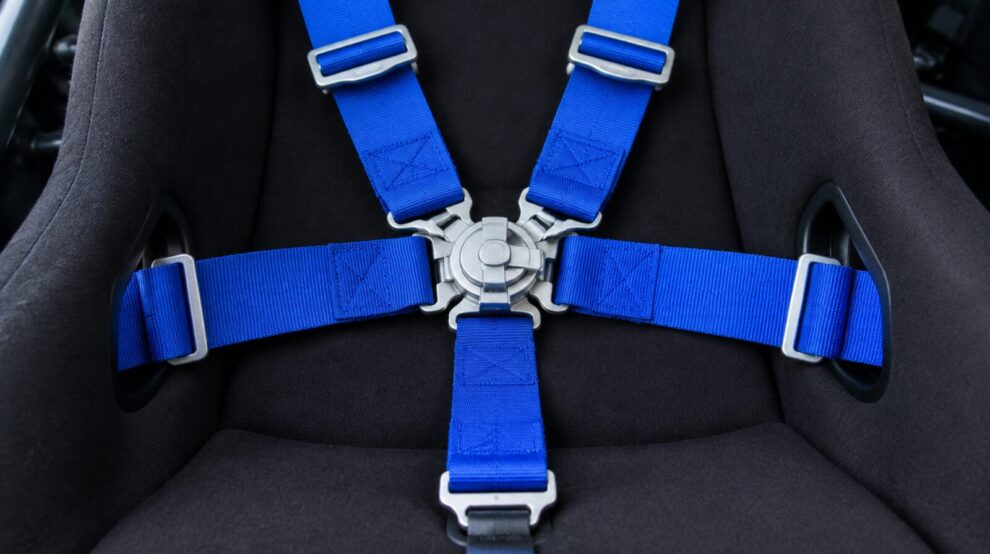 A racing harness with blue straps and a central camlock buckle across a black bucket seat