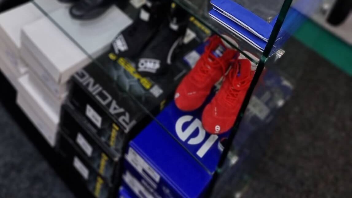 Race boots on the lower glass shelves of the GSM Performance showroom, with black OMP boots, red Sparco boots and boot boxes below