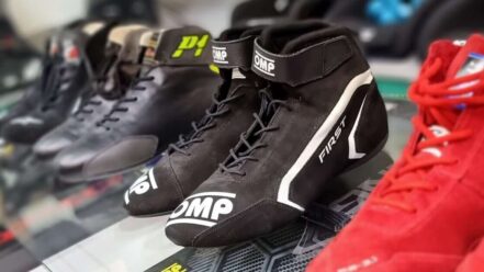 Black OMP First race boots on a glass display shelf beside a pair of red Sparco race boots, with racing seats behind