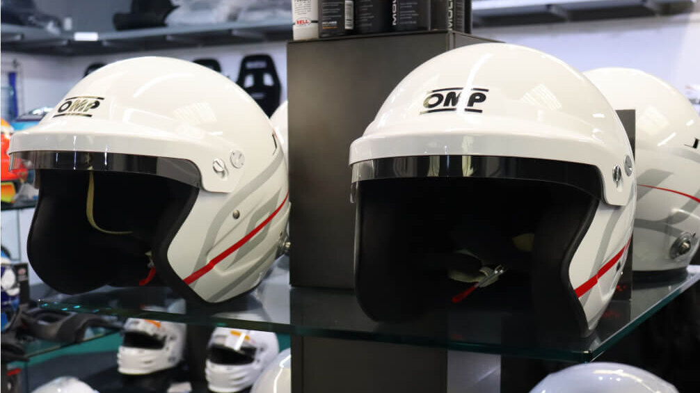 Two white OMP J-R open face helmets with red and grey stripes on a glass shelf in the GSM Performance showroom, with silver Stilo helmets below