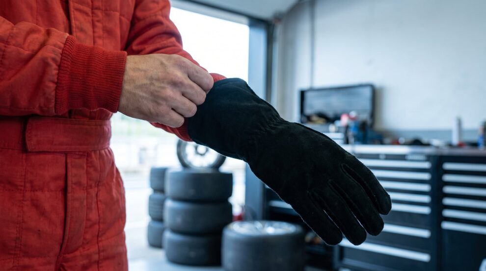 A driver in a red race suit in a pit garage pulling a black suede race glove on so the gauntlet goes over the knitted cuff of the suit sleeve