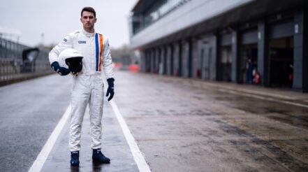 Driver in a white Sparco Gulf Replica race suit with a single blue and orange stripe down the chest, wearing navy Gulf Top race boots and navy Land race gloves, standing in a wet pit lane holding a white helmet