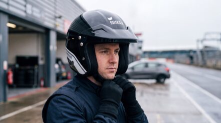 A driver in a plain navy race suit fastening the chin strap of a black and grey Sparco J-Pro open face helmet in a wet pit lane at a British circuit