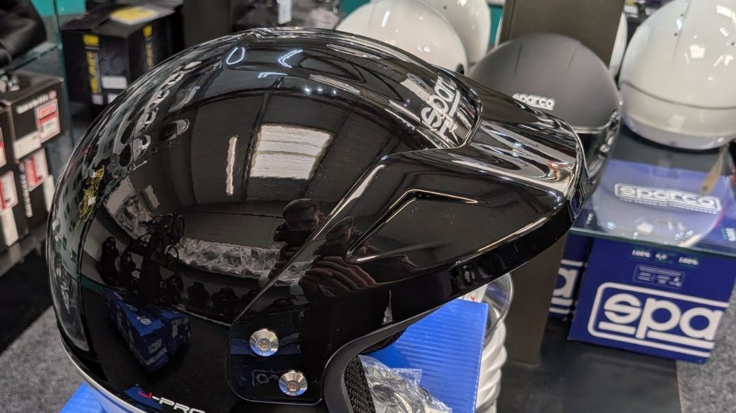 The black and grey Sparco J-Pro on its Sparco box in the showroom, the sparco wordmark across the crown, with white and matt black helmets on the shelf behind