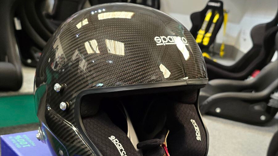 A Sparco RJ-i Carbon open face helmet showing its carbon weave, in the GSM Performance showroom with race seats behind