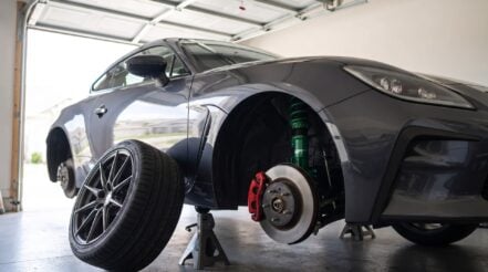 A green TEIN FLEX Z front coilover fitted in the wheel arch of a Toyota GR86 on axle stands