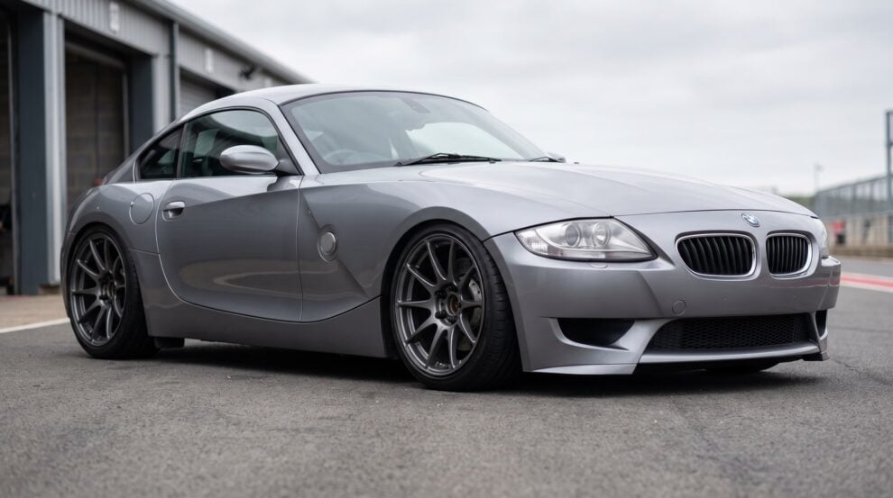 A BMW Z4 M Coupe in silver grey, sitting low on coilovers in a pit lane, dark aftermarket wheels with open hubs
