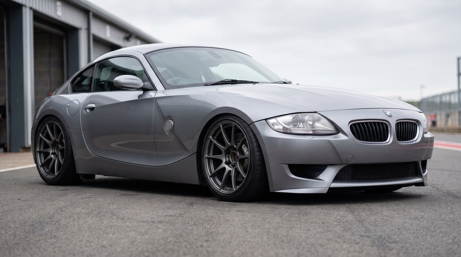 A BMW Z4 M Coupe in silver grey, sitting low on coilovers in a pit lane, dark aftermarket wheels with open hubs