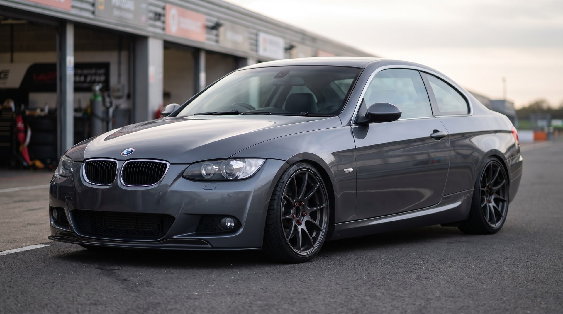 BMW E92 3 Series coupe on coilovers in a pit lane: arch gap closed, dark aftermarket wheels, a little camber
